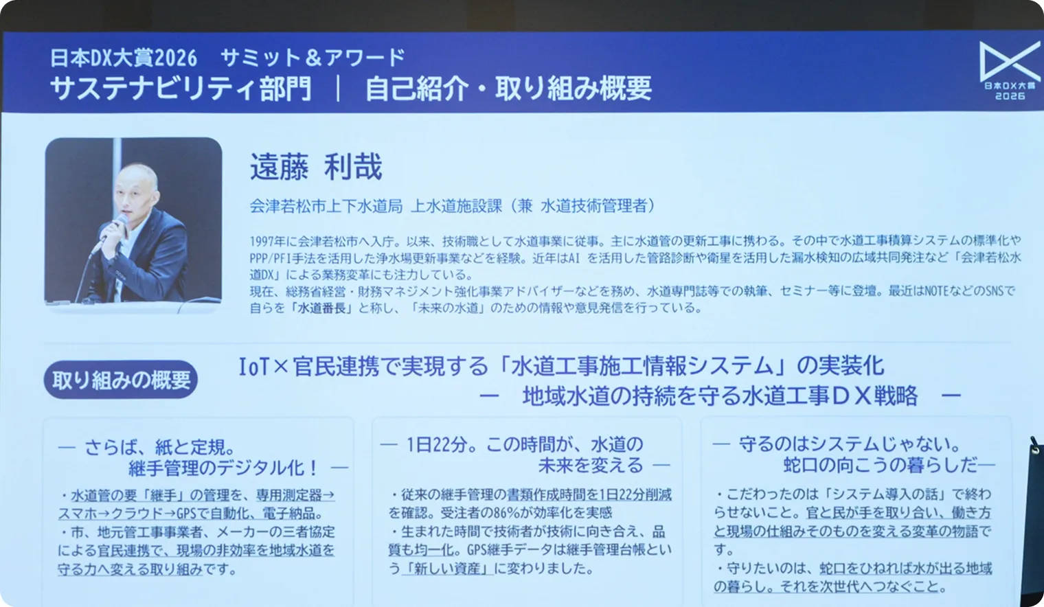 「日本DX大賞2026 サミット＆アワード」サステナビリティ部門のプレゼンテーションスライドを撮影した写真。会津若松市上下水道局 遠藤 利哉氏の自己紹介と、「水道工事施工情報システムの実装化 ― 地域水道の持続を守る水道工事DX戦略」の取り組み概要が示されている。手前には聴講者の姿。