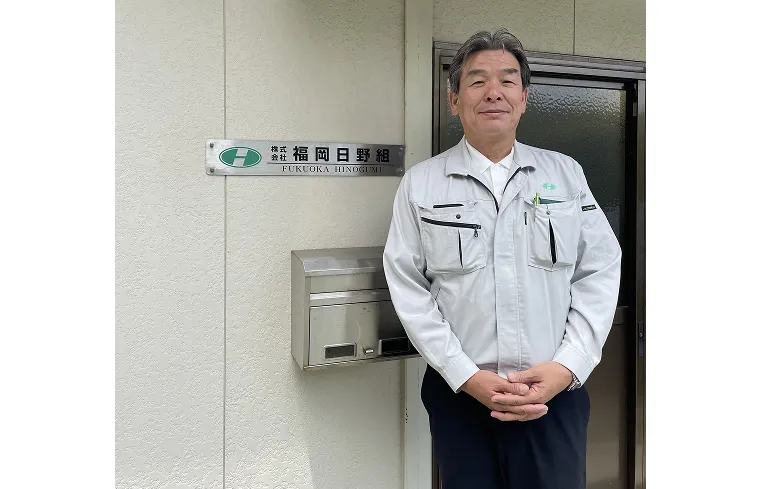 「株式会社 福岡日野組」の社名プレートの前に立つ、代表取締役社長 日野様。