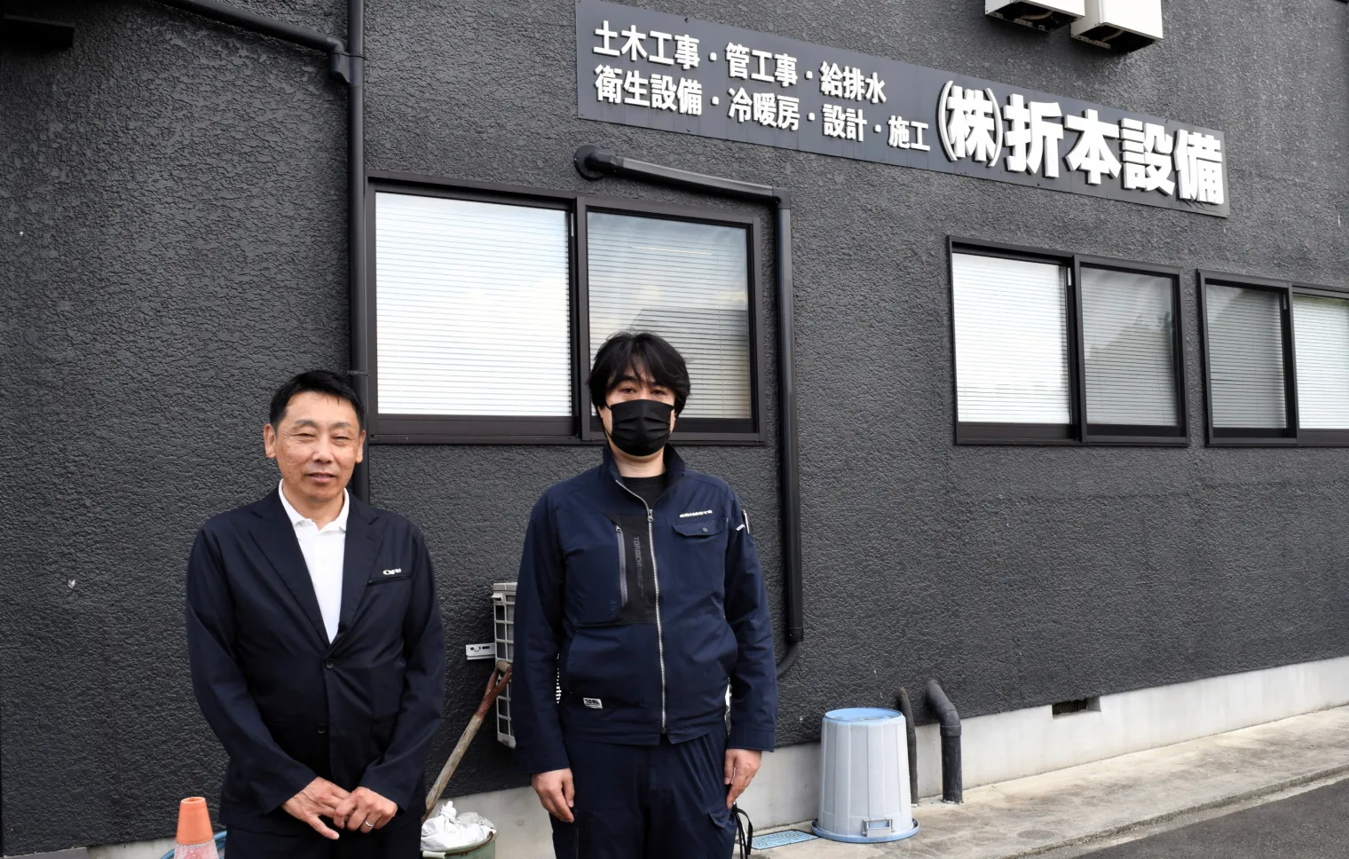 株式会社折本設備の社屋前に立つ折本社長と池田課長